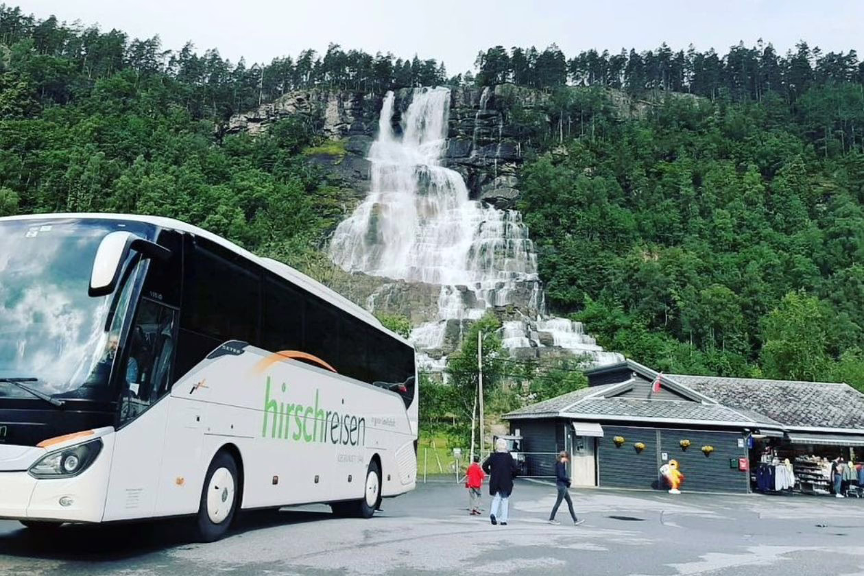 Bild einer Reise des Unternehmens Hirschreisen. Ein großer weißer Bus steht im Bild links, dahinter ist ein Wasserfall. Neben dem Bus laufen Reisende.