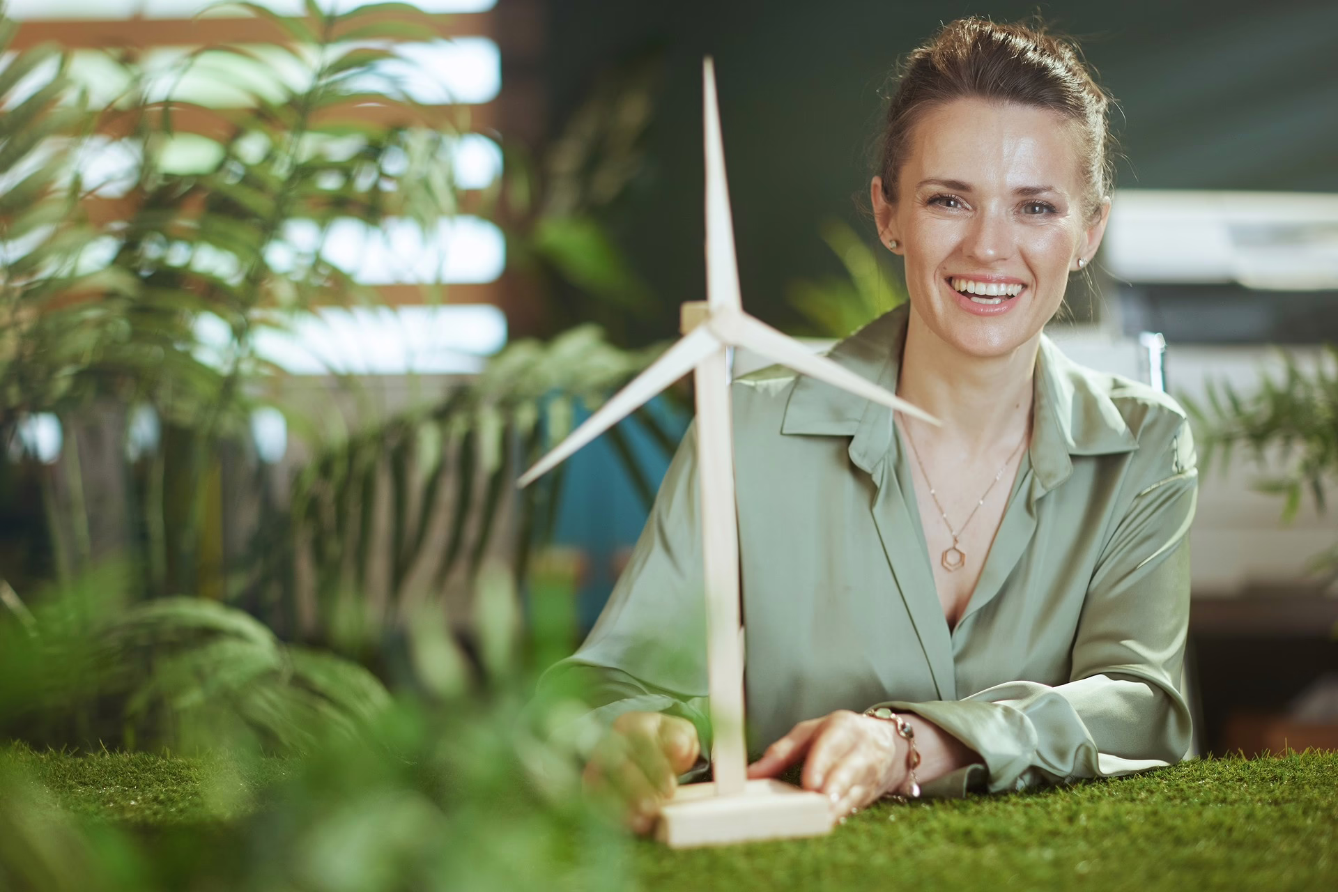 EIne Frau sitzt an einem Schreibtisch und lächelt und hält das Modell von einem Windrad in die Kamera. Sie trägt eine Bluse.